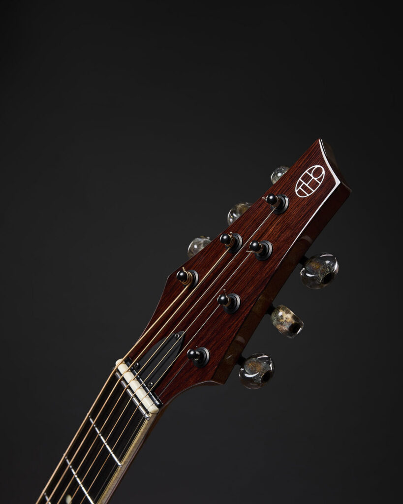 Close-up of Thom Orgler Guitars headstock with inlay logo and tuning machines on a handcrafted acoustic guitar, dark studio background.